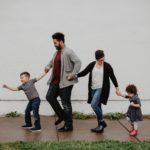 Four people are holding hands and walking down the sidewalk. A young child in a grey t-shirt is in the lead, pulling the rest of the group forward. Behind him is a grown-up in a grey cardigan and red shirt, followed by a shorter adult in a black cardigan and white shirt. A smaller child in a black dress is at the tail end of the group. The backdrop is a white building.