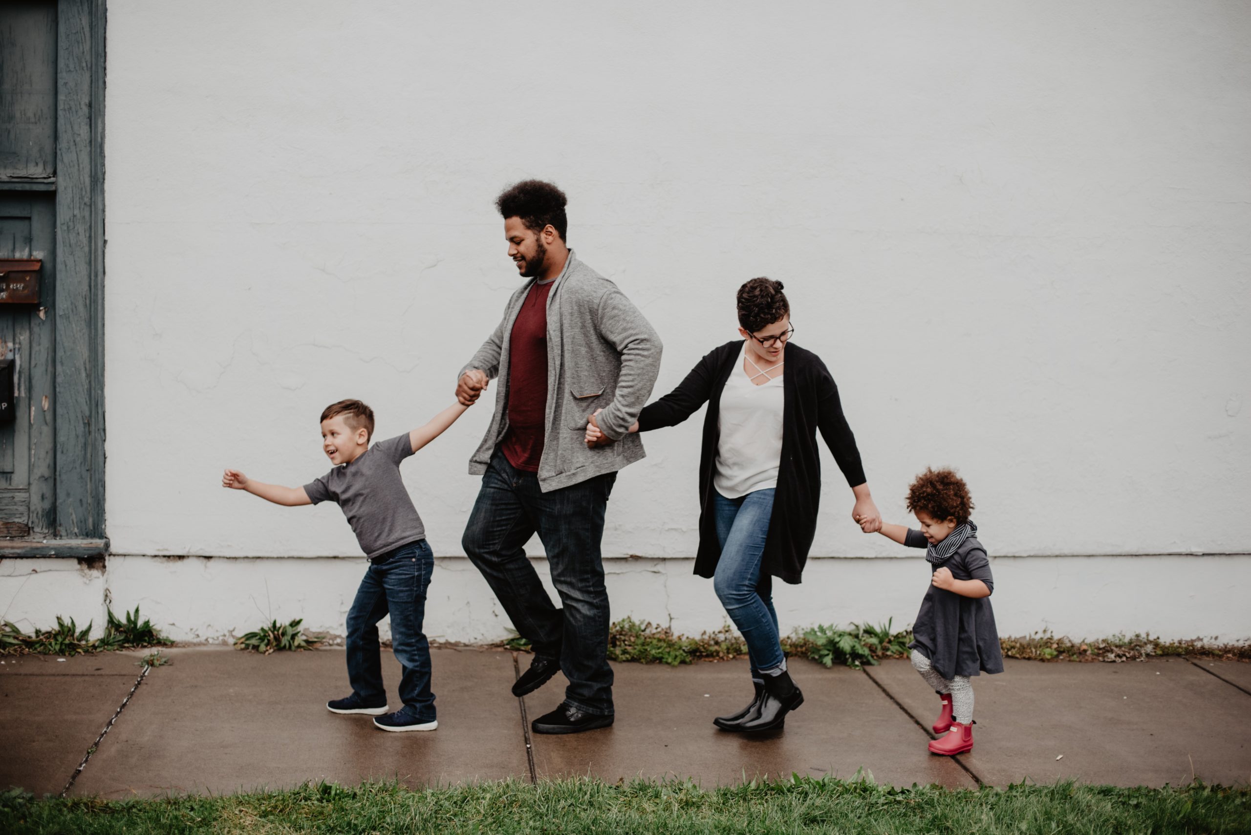 Four people are holding hands and walking down the sidewalk. A young child in a grey t-shirt is in the lead, pulling the rest of the group forward. Behind him is a grown-up in a grey cardigan and red shirt, followed by a shorter adult in a black cardigan and white shirt. A smaller child in a black dress is at the tail end of the group. The backdrop is a white building.